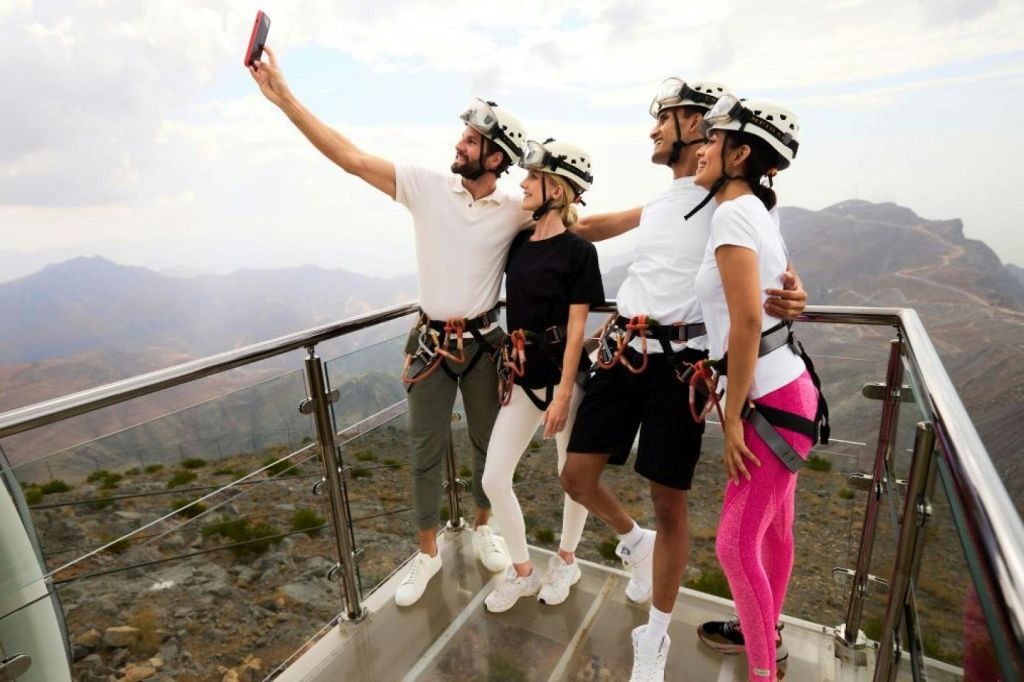 산이 내려다보이는 유리 플랫폼에서 헬멧과 마구를 착용한 네 명이 셀카를 찍고 있습니다.