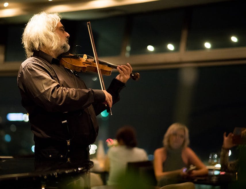 한 사람이 밤에 실내에서 바이올린을 연주하고, 배경에 앉은 후원자들이 바이올린을 관찰하고 식사를 합니다.
