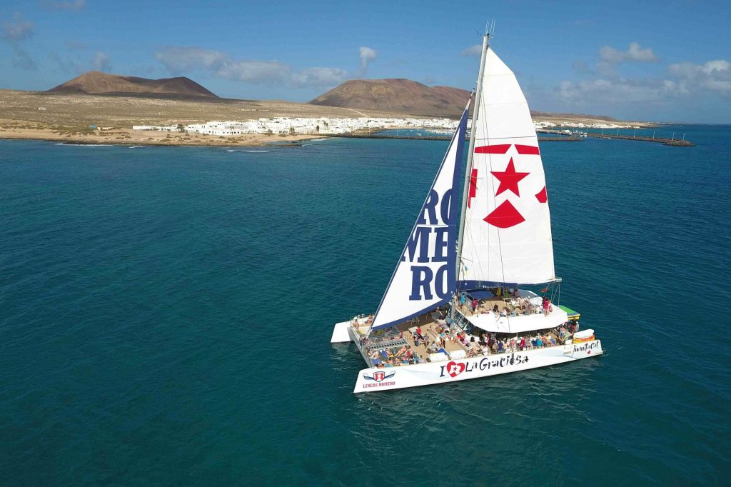 "I ❤️ La Graciosa" 가 있는 쌍동선과 돛단 항해, 하얀 건물과 언덕이 있는 해안 마을 근처의 푸른 물에서 항해.