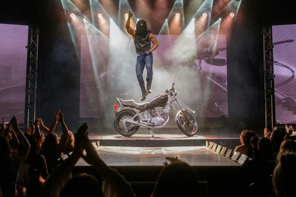 헬멧과 가죽 조끼를 쓴 공연자가 무대에서 오토바이 위로 공중으로 뛰어오르고, 관객이 박수를 치고 있습니다.