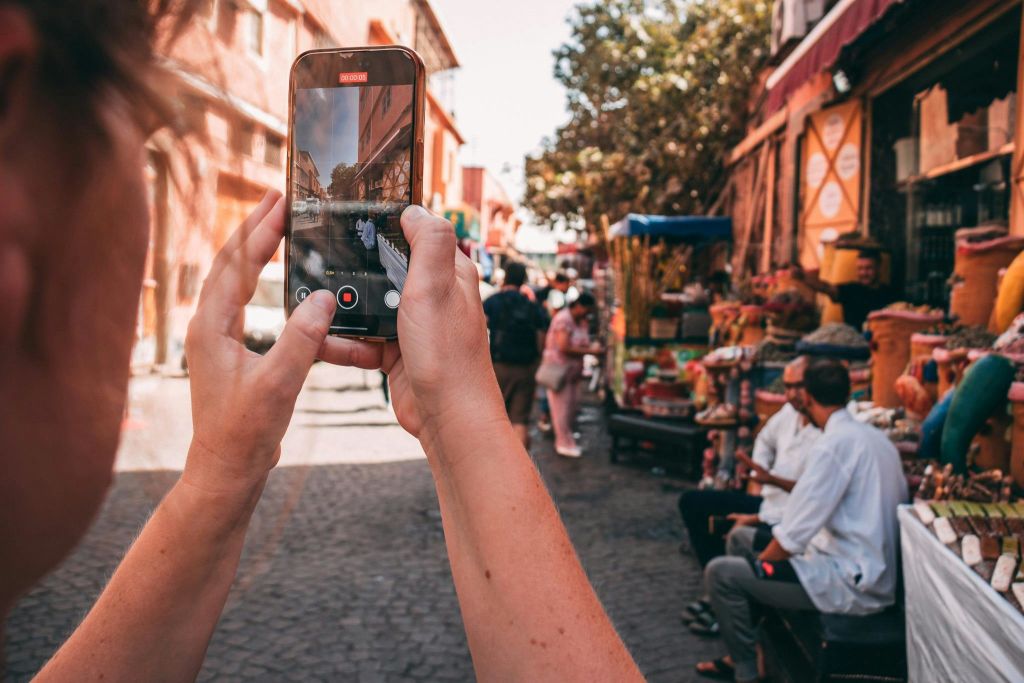 스마트폰을 들고 사람들, 화려한 가판대, 햇살이 비치는 건물이 있는 번잡한 야외 시장을 촬영하는 사람.