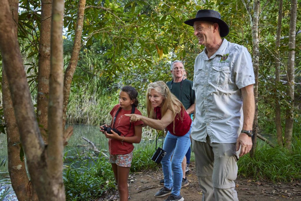 쌍안경을 든 소녀와 가리키는 여성, 두 남자가 물 근처 숲의 흙길에 서 있습니다.