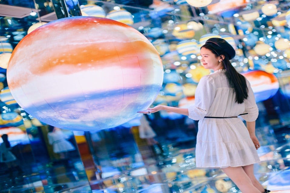 흰색 드레스와 베레모를 입은 여성이 반사되고 밝게 빛나는 공간에서 커다랗고 화려한 구형 물체 근처에서 포즈를 취하고 있습니다.