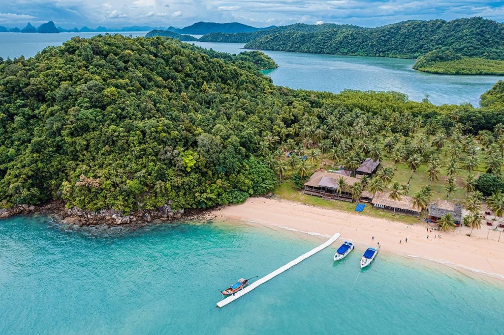 부두에 정박한 보트, 빽빽한 푸른 숲, 해안가 근처의 건물이 있는 열대 해변의 공중 전망.