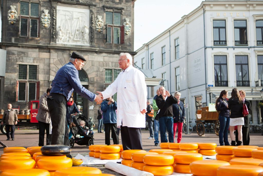 전경과 건물 뒤에 노란색 치즈 바퀴가 쌓여 있는 시장에서 두 남자가 악수하고 있습니다.