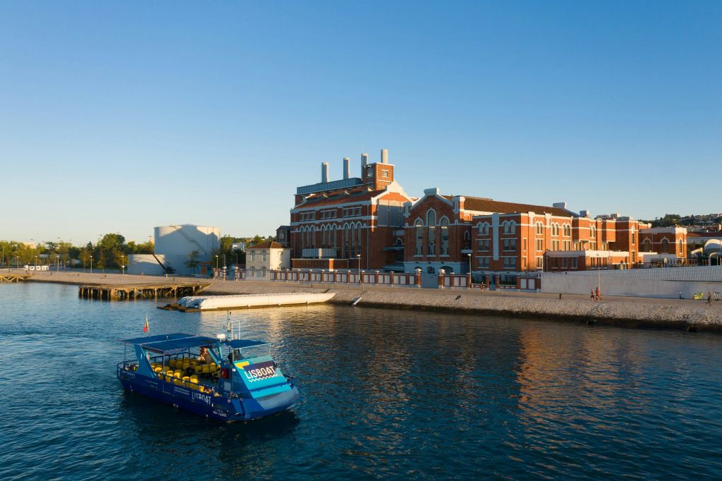 "USBOAT" 가 프린트된 파란색 투어 보트가 맑고 푸른 하늘 아래 물가의 크고 유서 깊은 붉은 벽돌 건물을 지나고 있습니다.