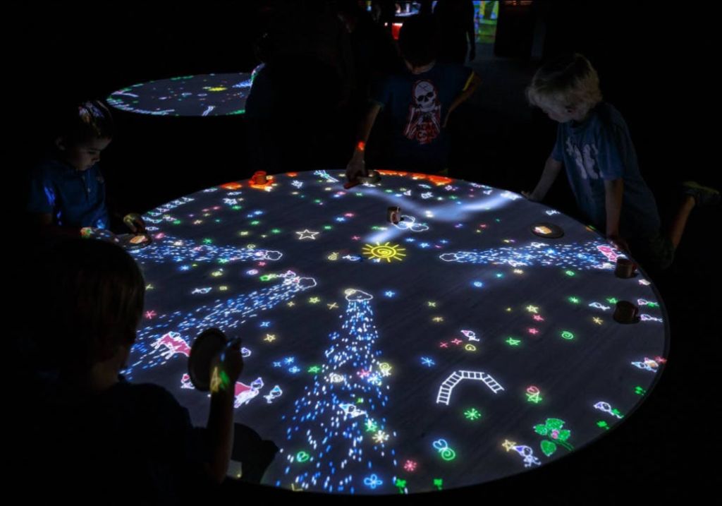 어두운 방에서 다채로운 애니메이션 그래픽을 표시하는 둥근 조명 테이블과 상호 작용하는 어린이.