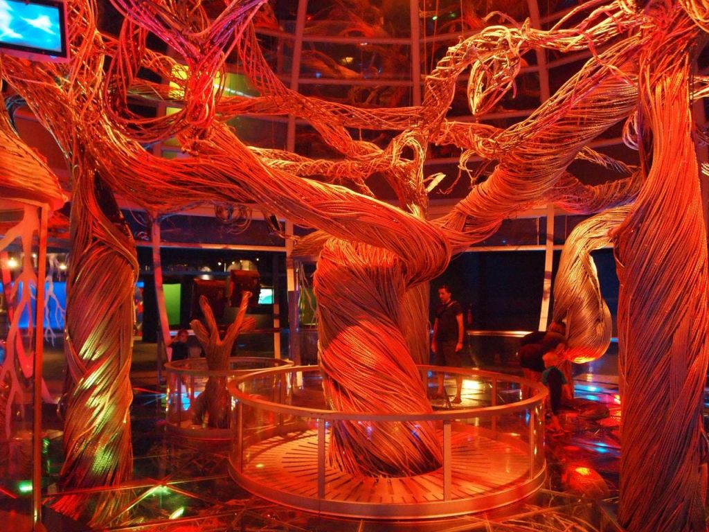 빨간색과 주황색 조명으로 비춰진 직조된 재료로 만든 복잡한 나무 조각품으로, 몇 명이 관찰하고 있습니다.