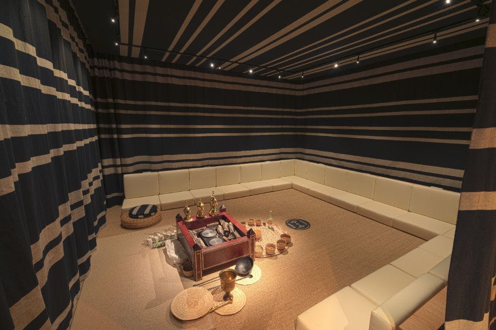 파란색 줄무늬 벽과 천장, 흰색 L자형 좌석, 물품이 놓인 중앙 트레이, 바닥에 직조된 바구니와 매트가 있는 객실입니다.