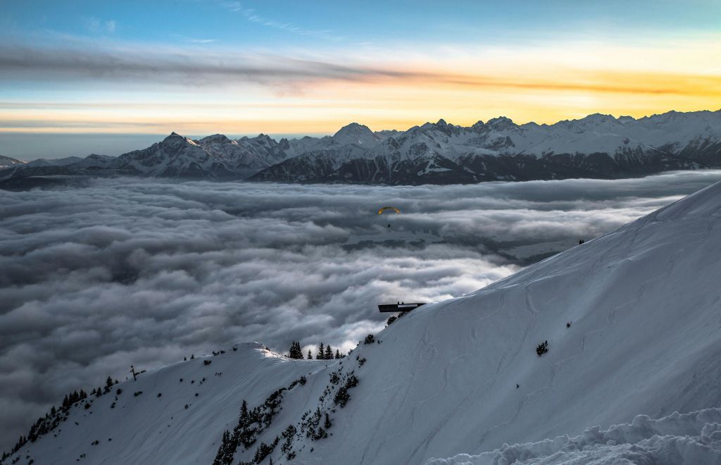 눈 덮인 산비탈 아래 구름 바다와 해가 뜨는 먼 산이 내려다보이는 나무 벤치가 있습니다.