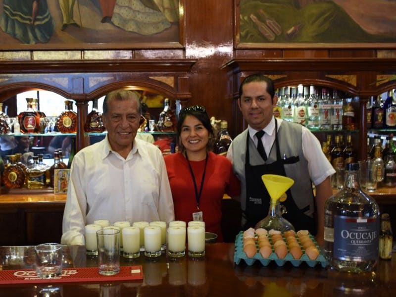 세 사람이 바 뒤에 서서 미소를 짓고 있습니다. 바 카운터에는 여러 음료와 달걀 쟁반이 있습니다. 병이 구비된 선반이 그 뒤에 있습니다.