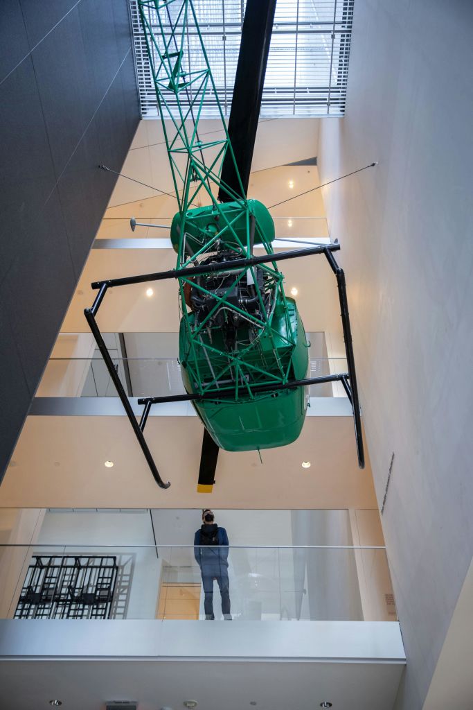 한 사람이 낮은 층에 서서 현대식 건물 아트리움의 천장에 매달린 녹색 헬리콥터를 관찰하고 있습니다.