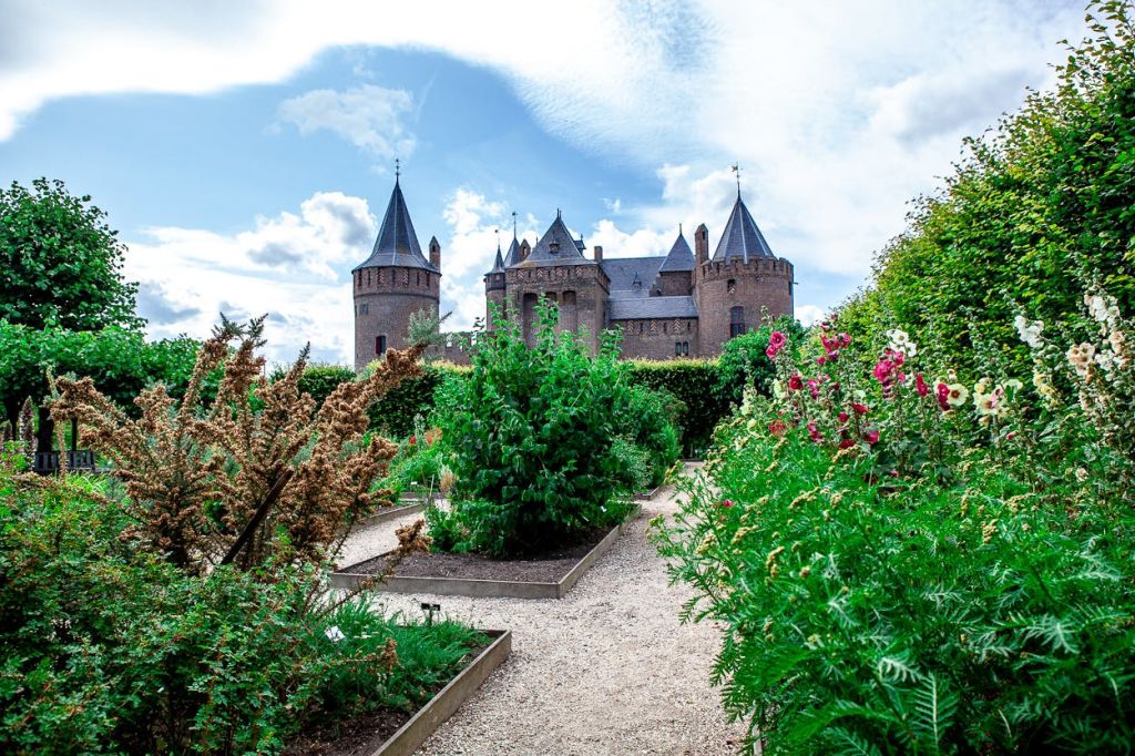 부분적으로 흐린 하늘 아래 다양한 식물과 꽃이 가득한 정원에서 여러 개의 탑이 보이는 커다란 벽돌 성.