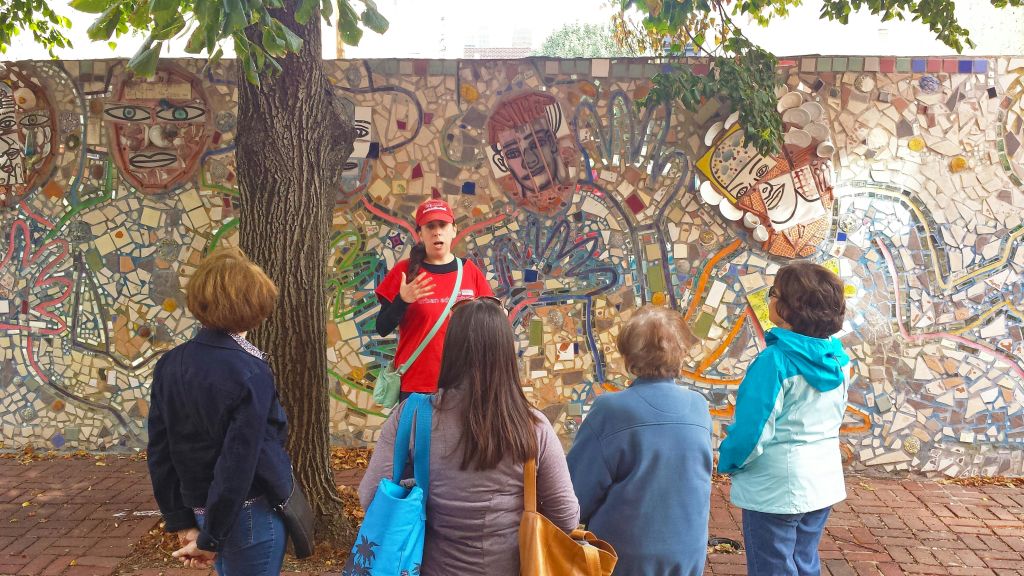 빨간 모자와 셔츠를 입은 가이드가 나무 아래 화려한 모자이크 벽 앞에서 네 사람에게 말을 걸고 있습니다.