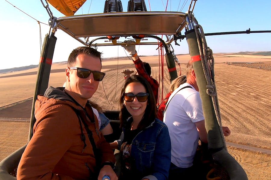 광활한 사막 풍경 위에 떠 있는 열기구 바구니 안에서 선글라스를 쓴 두 사람이 미소를 짓고 있습니다.