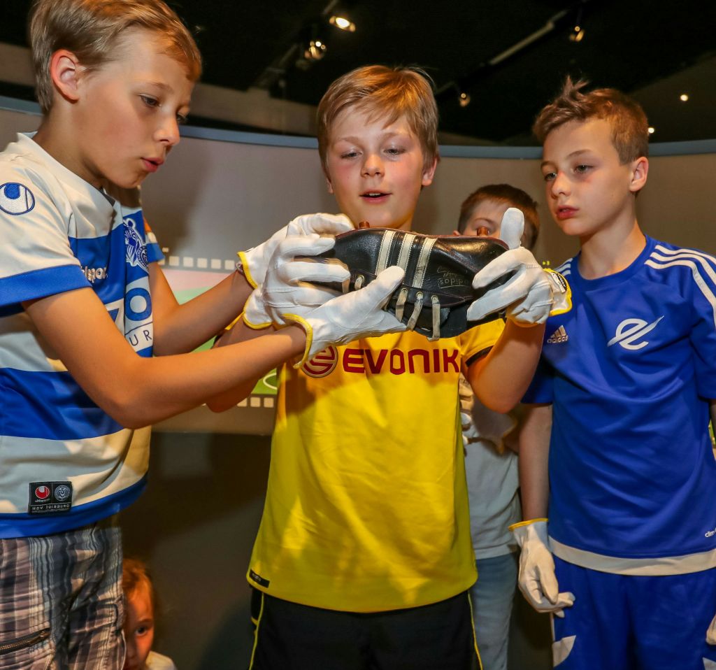 축구 유니폼과 흰색 장갑을 착용한 세 명의 소년이 박물관과 같은 분위기에서 신발을 살펴보고 있습니다.