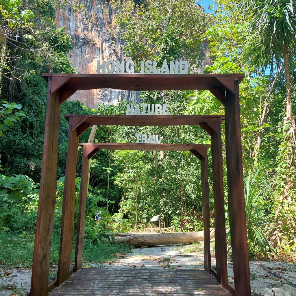울창한 녹음과 바위 절벽으로 둘러싸인 "홍섬 자연 산책로" 표지판이 있는 나무 아치길.