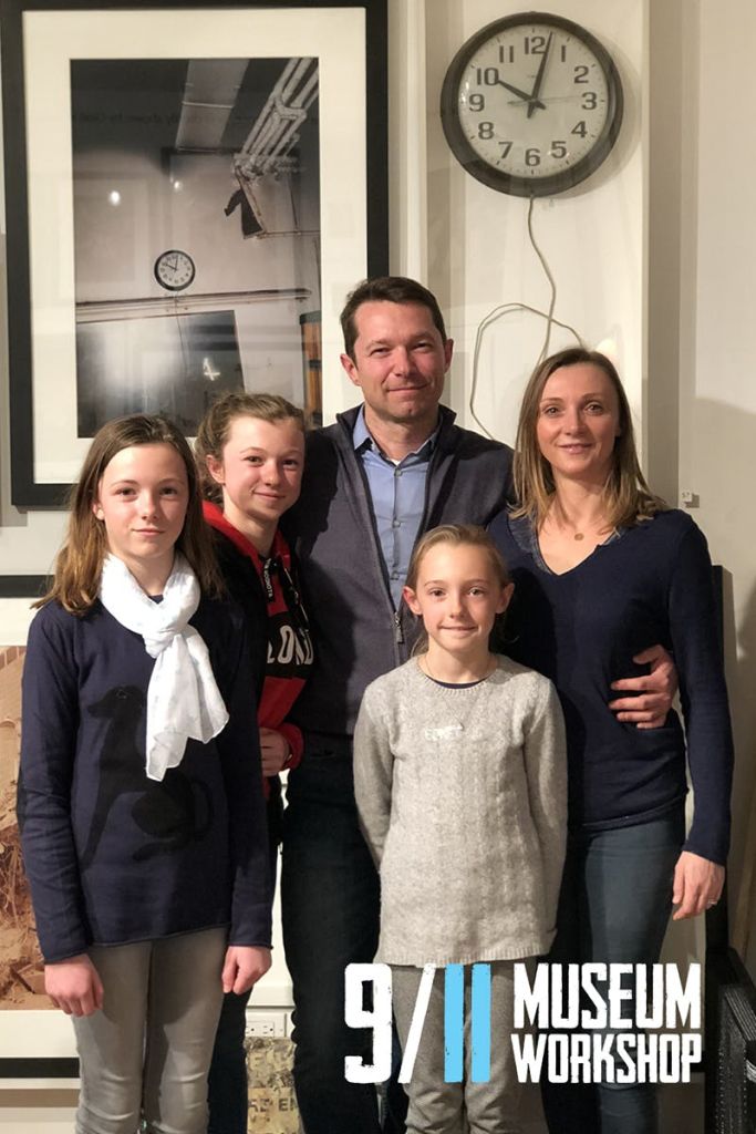 5인 가족이 함께 실내에 서서 미소를 짓고 있습니다. 시계와 액자 그림이 벽에 보입니다.