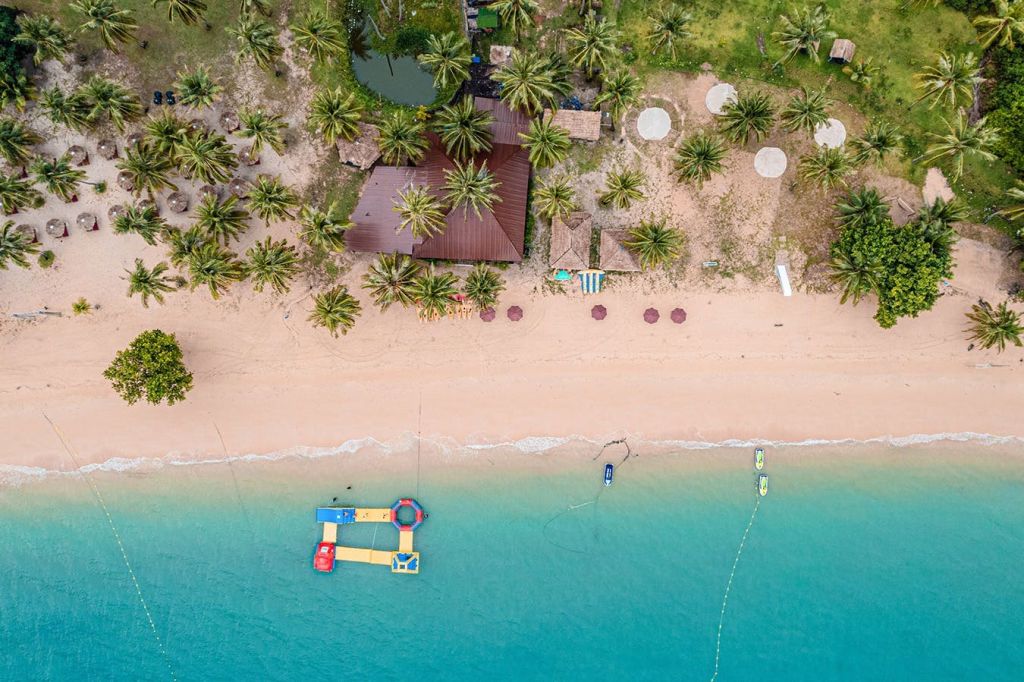 청록색 물, 비치 파라솔, 야자수, 녹음으로 둘러싸인 목조 건물이 있는 해변의 공중 전망.