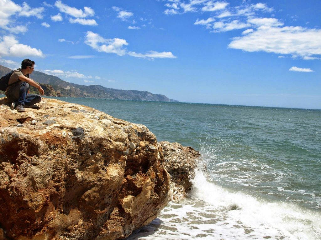 바다 옆의 큰 바위에 앉아 파도가 부서지는 사람. 구름이 낀 맑은 하늘과 먼 산이 보입니다.