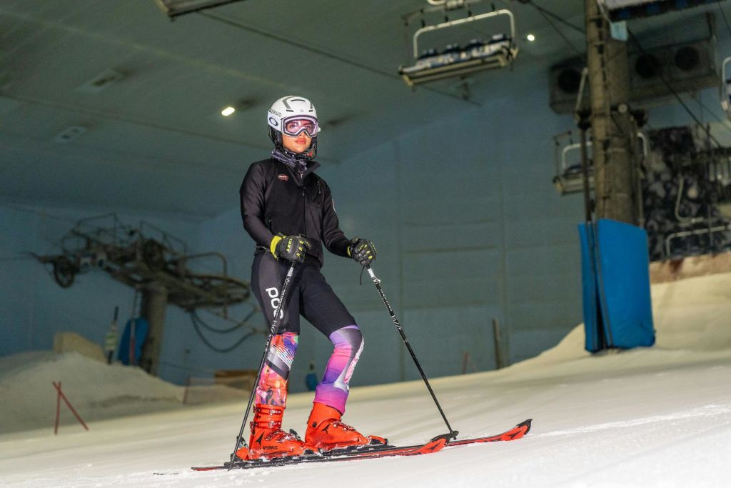 스키 장비를 착용한 사람이 실내 스키 슬로프에 서서 스키 폴을 잡고 있으며, 스키 리프트를 배경으로 서 있습니다.