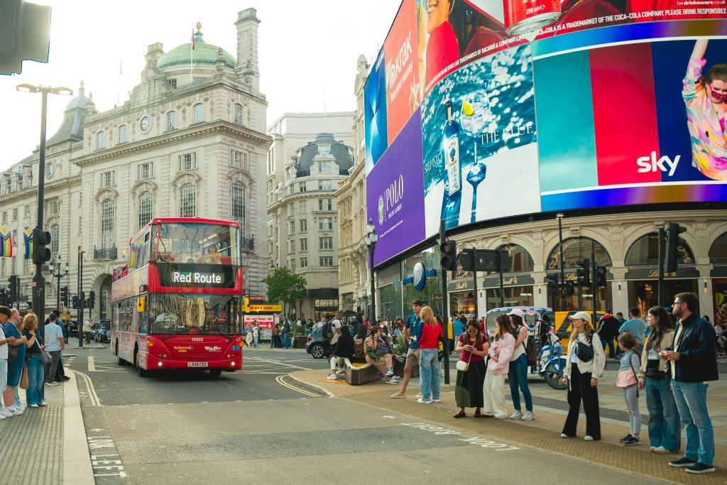 보행자 그룹 근처의 도시 거리에 있는 빨간색 이층 버스와 대형 전자 광고판이 있는 건물.