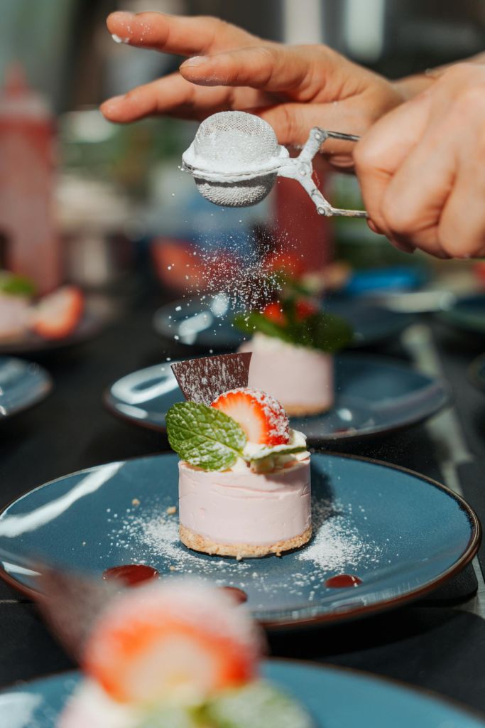 파란색 접시에 민트 잎으로 장식된 딸기 치즈케이크에 손으로 가루 설탕을 뿌립니다.