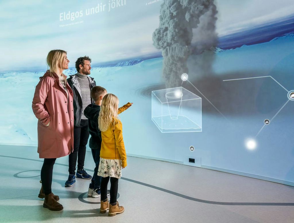 네 명의 가족이 빙하 아래에서 화산 폭발을 보여주는 대화형 전시물을 관찰하고 텍스트와 다이어그램을 표시합니다.