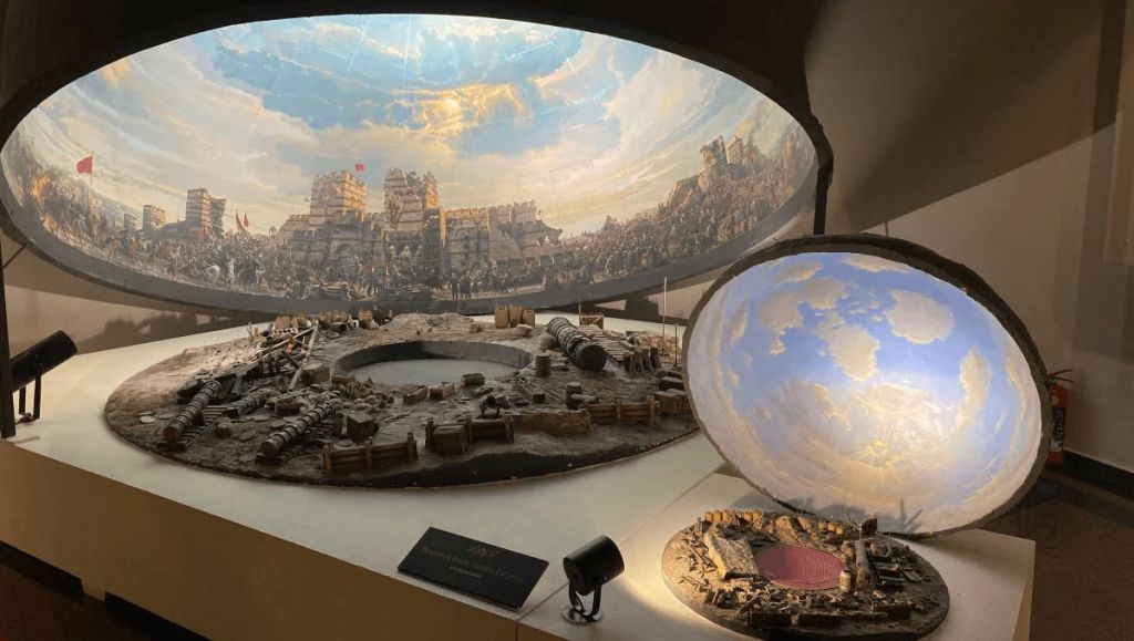 박물관에 전시된 포위된 고대 도시의 디오라마에는 미니어처 건물, 포위 장비, 하늘을 보여주는 부분 돔이 완비되어 있습니다.