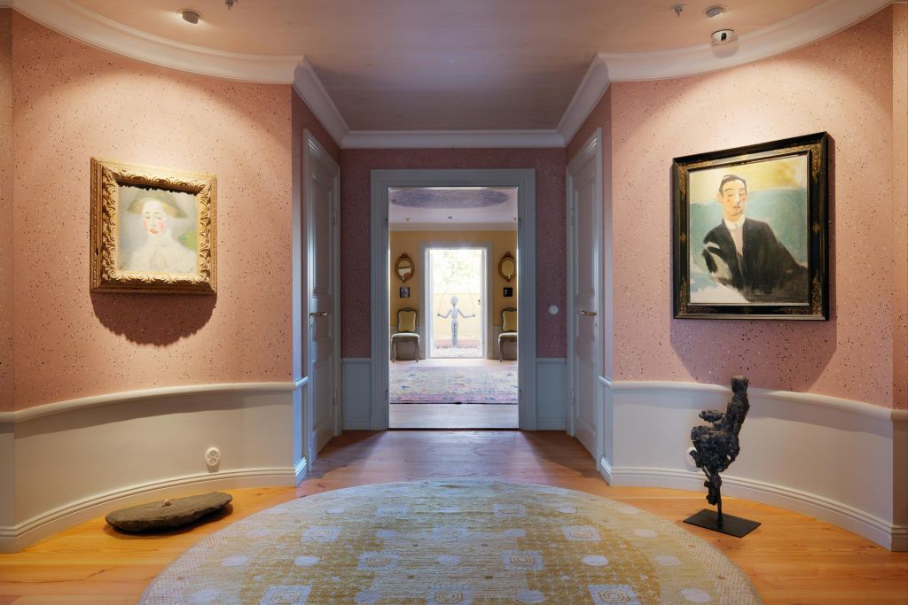 분홍색 벽에 초상화 그림이 그려진 복도, 받침대에 앉은 조각상, 원형의 노란색 무늬 러그가 특징입니다.