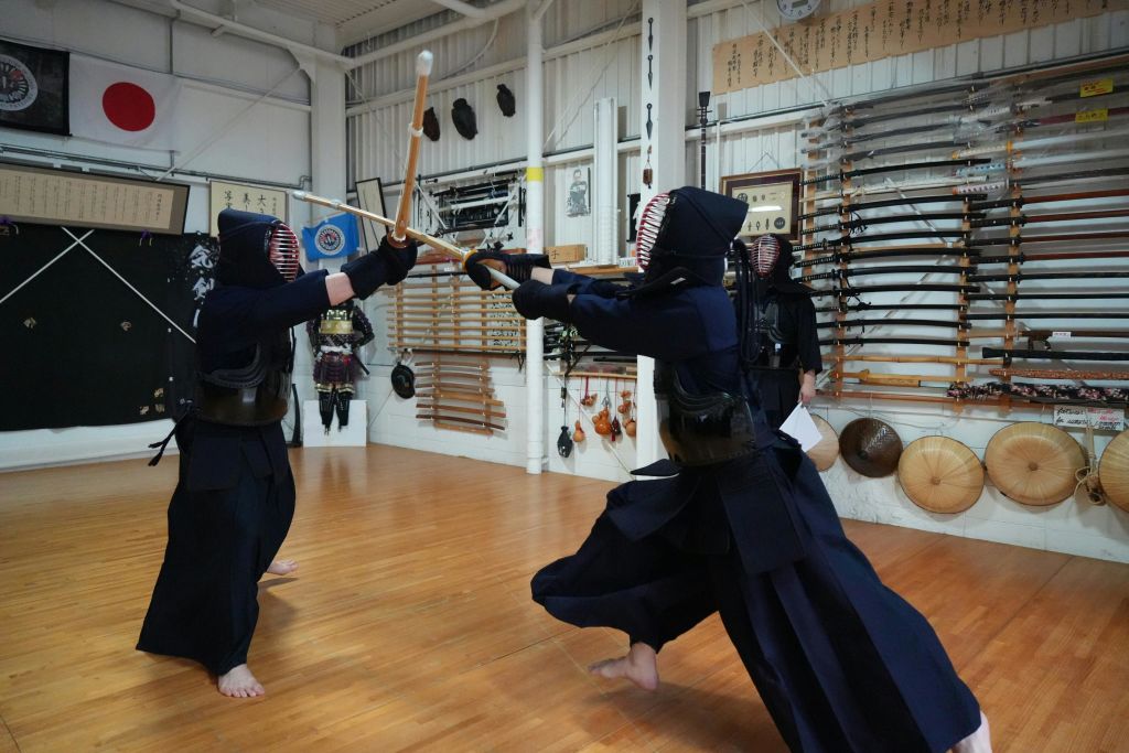 전통 검도를 입은 두 사람이 도장에서 대나무 검을 들고 벽에 다양한 무술 장비를 걸치고 스파링을 합니다.