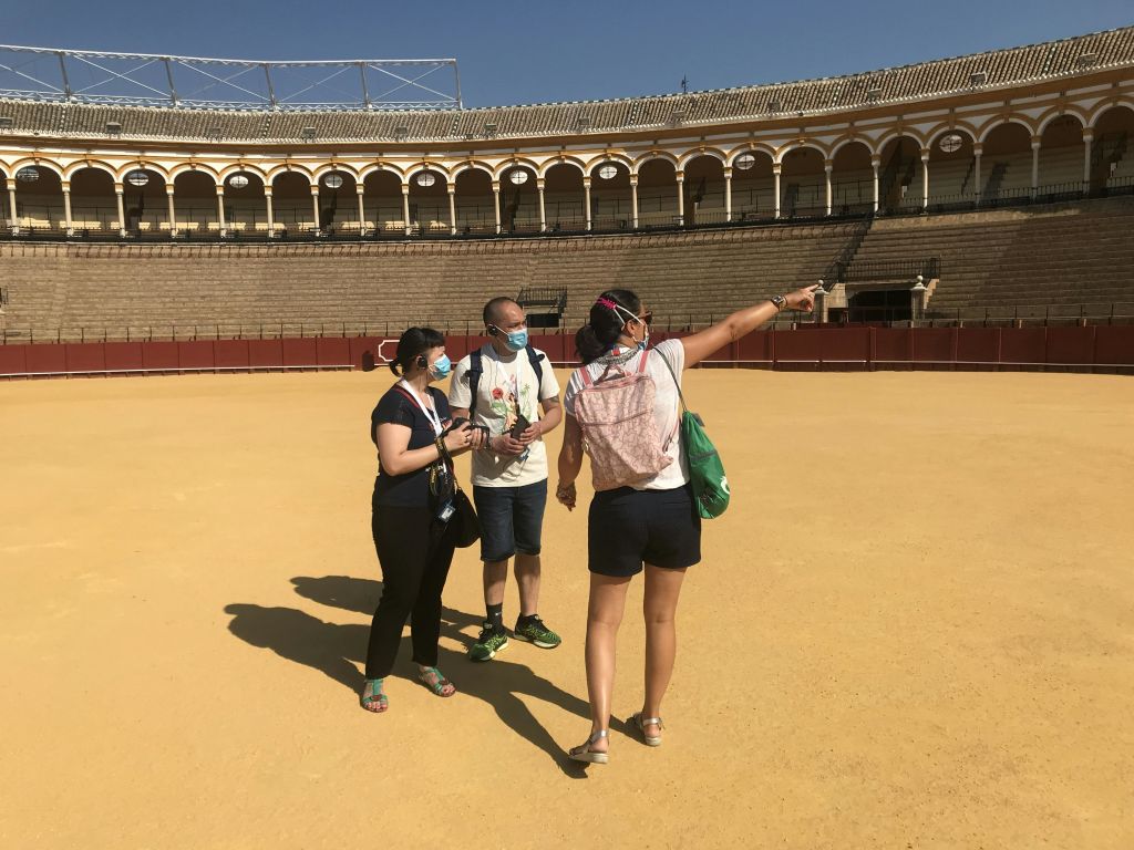 안면 마스크를 쓴 세 사람이 아치형 좌석 공간이 있는 커다랗고 텅 빈 황소 고리에 서 있습니다. 한 사람이 가리키고 있습니다.