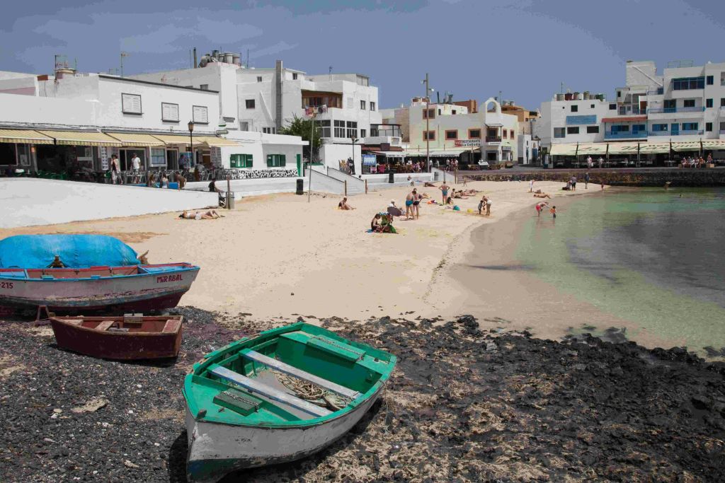 일광욕과 수영을 즐기는 사람들이 있는 작은 해변으로, 흰색 건물 옆에 있습니다. 전경의 바위 해안에 두 척의 노젓는 배가 있습니다.