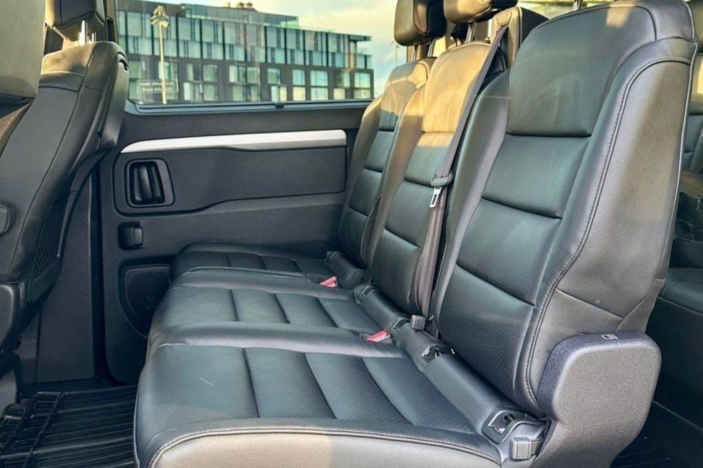 검은색 가죽 시트 3개가 있는 차량의 뒷좌석 모습. 햇살이 비추면 창문을 통해 현대식 건물이 보입니다.