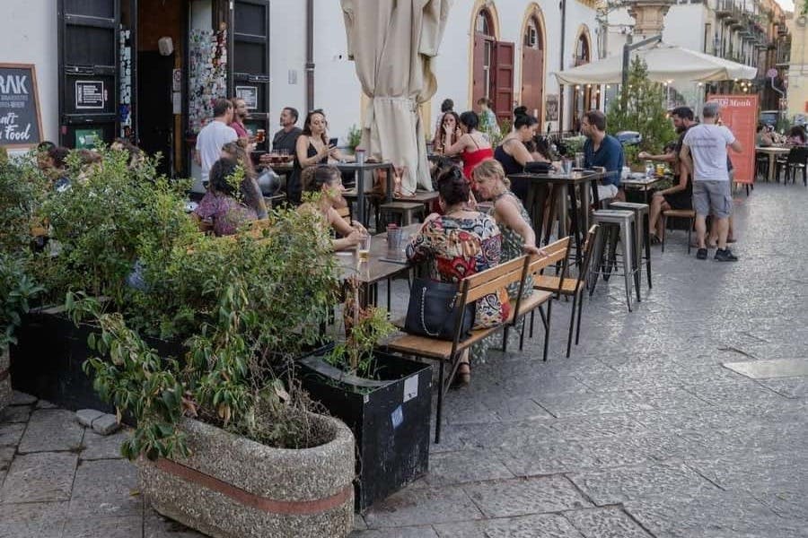 화분과 커다란 우산이 놓인 카페 야외 테이블에서 식사와 대화를 즐기는 사람들.