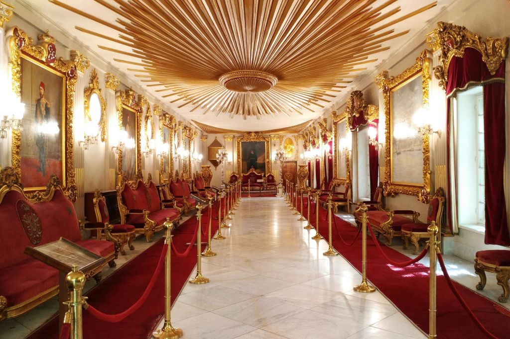 레드 카펫, 골드 프레임 거울, 샹들리에, 레드 벨벳 의자, 황금색 받침대, 정교한 천장 디자인이 돋보이는 화려한 홀.