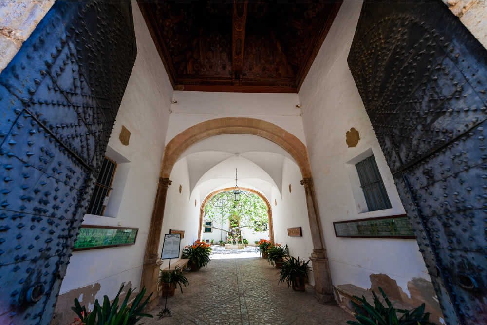 화분과 샹들리에로 장식된 아치형 천장이 있는 하얀 벽의 복도는 햇살이 내리쬐는 안뜰로 이어집니다.