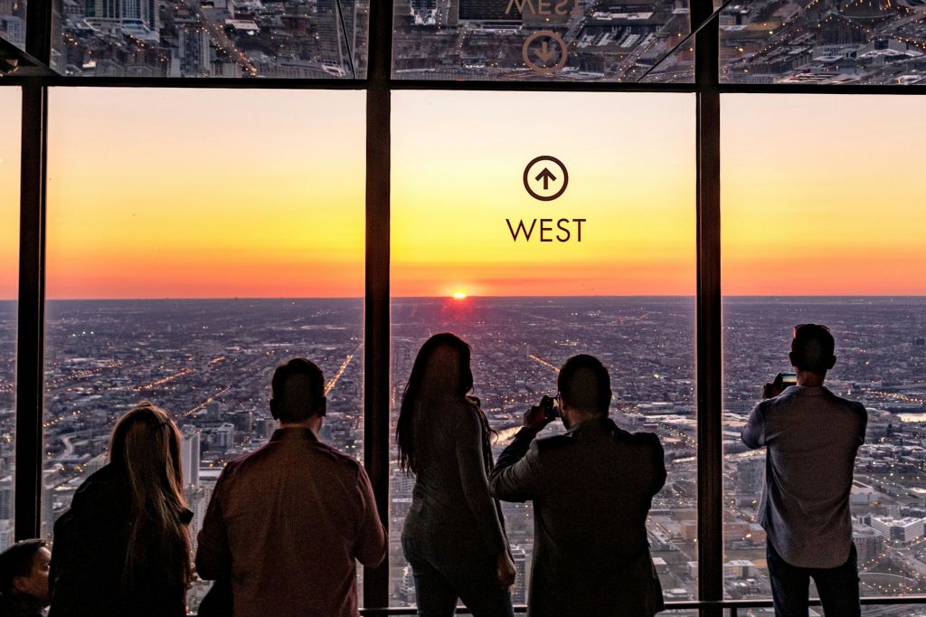 고층 건물 안에 네 사람이 서서 '서쪽' 으로 표시된 커다란 창문을 통해 도시 풍경의 일몰을 감상하고 있습니다.