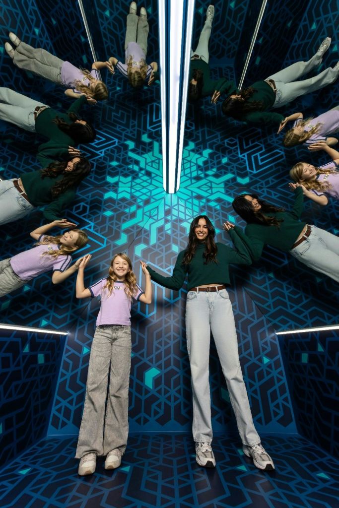 기하학적 파란색 패턴이 있는 거울 방에 두 사람이 서서 주변을 무한히 반사합니다.