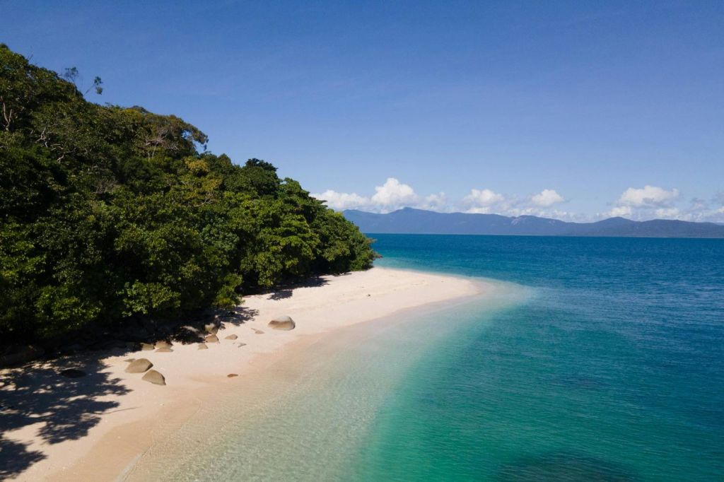 하얀 모래사장과 맑은 청록색 바다, 커다란 바위, 푸른 하늘 아래 푸른 나무가 우거진 한적한 해변.