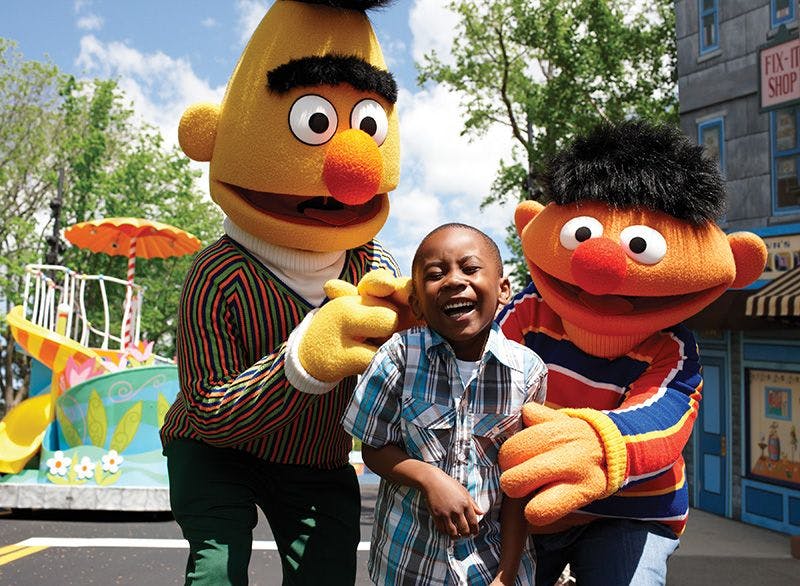 화창한 날 야외에서 커다란 노란색 꼭두각시와 주황색 꼭두각시 옷을 입은 두 명의 캐릭터와 함께 미소 짓고 포즈를 취하는 아이.