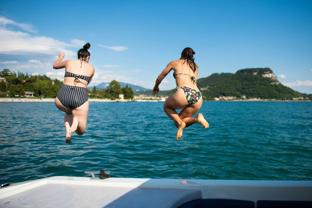 수영복을 입은 두 명의 여성이 배를 타고 호수로 뛰어들고, 언덕과 멀리 떨어진 건물이 맑고 푸른 하늘을 배경으로 펼쳐집니다.