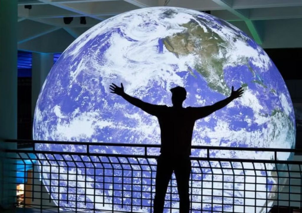 현대적인 전시 공간에서 커다란 지구 전광판 앞에 팔을 뻗은 사람의 실루엣.