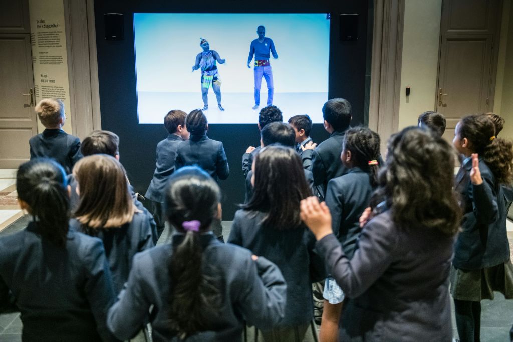교복을 입은 한 무리의 어린이들이 두 사람이 춤을 추는 화면을 보고 있습니다.
