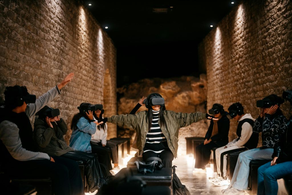 VR 헤드셋을 착용한 한 무리의 사람들이 돌담이 있는 방의 벤치에 앉아 있으며, 일부는 팔을 쭉 뻗고 가상 현실을 경험합니다.