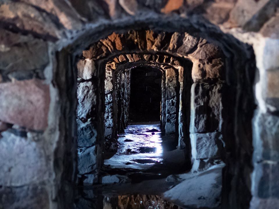 어두운 조명, 거친 돌담, 바닥에 물 반사가 있는 고대 석조 아치형 복도.