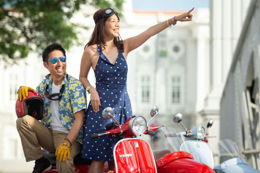 빨간색 전동 스쿠터를 탄 쾌활한 남녀가 건물과 나무를 배경으로 저 멀리를 가리키고 있습니다.