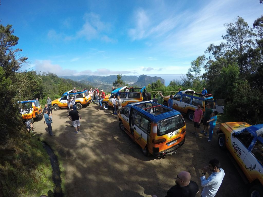 한 무리의 사람들이 산과 숲을 배경으로 흙길에 주차된 여러 대의 오프로드 차량 근처에 모였습니다.