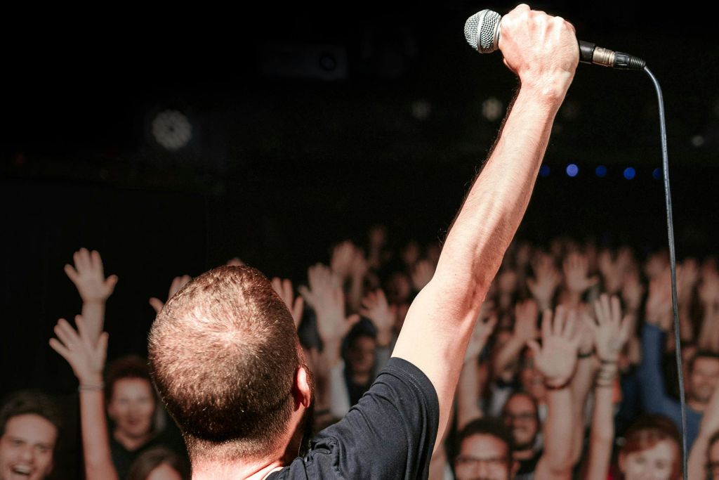 무대 위의 한 사람이 공중에서 마이크를 잡고, 수많은 사람들이 뒤에서 손을 들고 있습니다.
