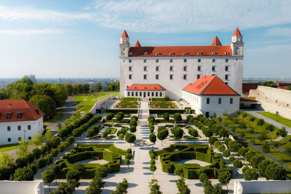 붉은 지붕과 여러 개의 탑이 있는 성은 기하학적 울타리와 가로수 통로가 있는 잘 다듬어진 정원을 내려다보고 있습니다.